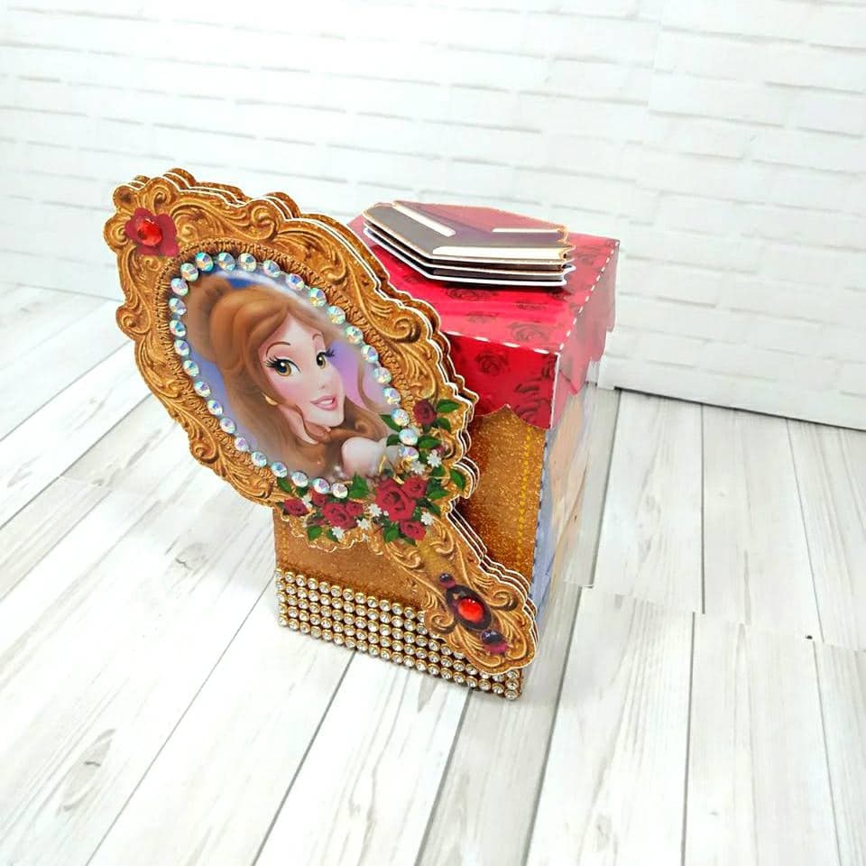 Caixa do tema Bela e a Fera com aplique em formato de espelho de mão, retrato oval de Bela emoldurado em dourado com pedraria e uma pequena rosa vermelha, tampa estampada com rosas, base glitter dourada e um livrinho de papel dobrado no topo