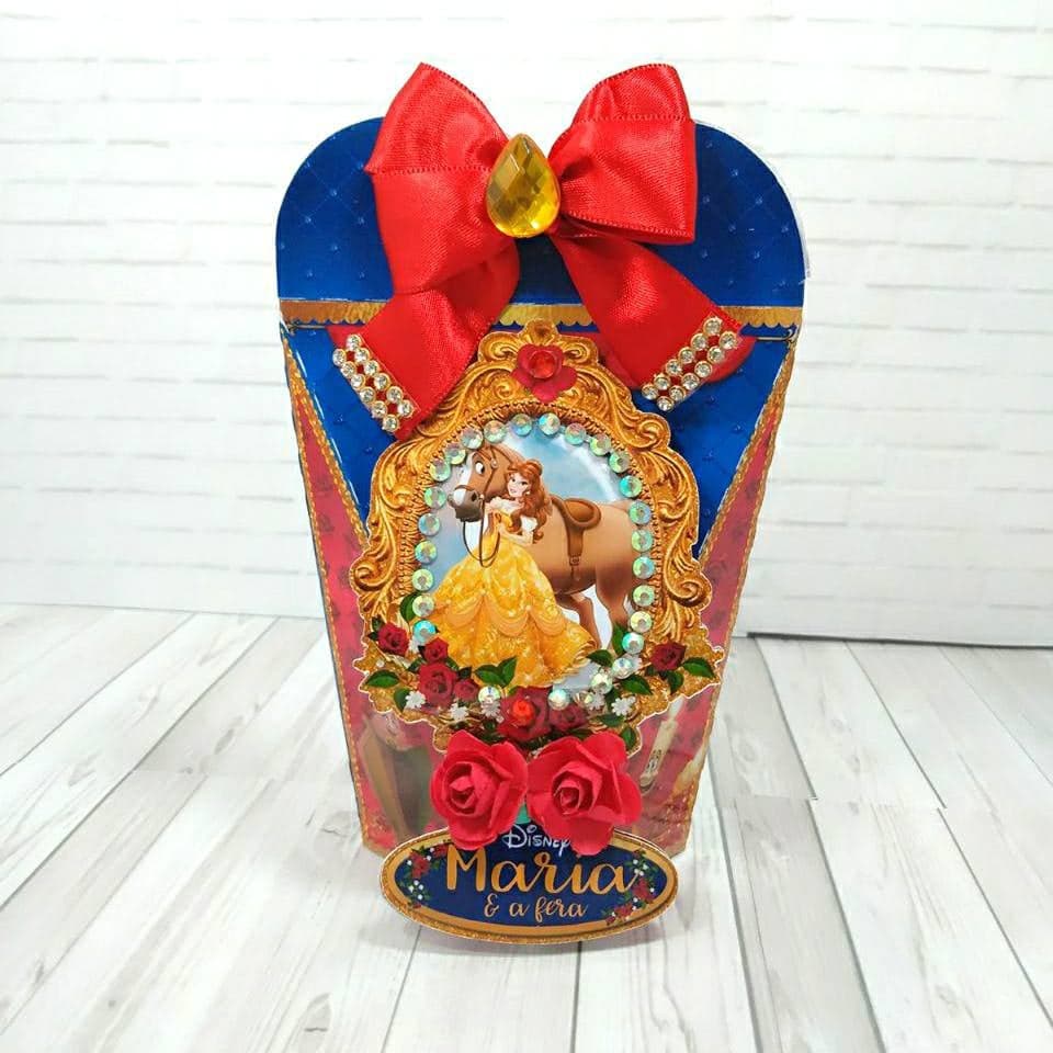 Caixa de presente arredondada amarrada com laço grande de cetim vermelho com gema facetada âmbar no centro, retrato oval de Bela com um cavalo emoldurado em dourado com pedraria, rosas de papel e borda de pedraria abaixo