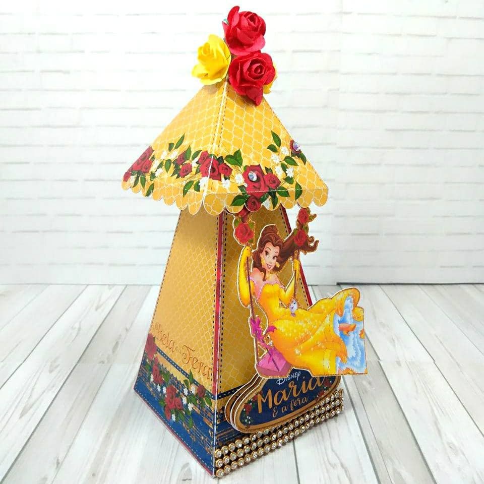 Caixa em formato de tenda com telhado piramidal em padrão dourado quadrifólio bordado com grinalda de rosas vermelhas, coroada por rosas de papel 3D vermelhas e amarelas, recorte de Bela em um balanço floral na frente e borda de pedraria na base