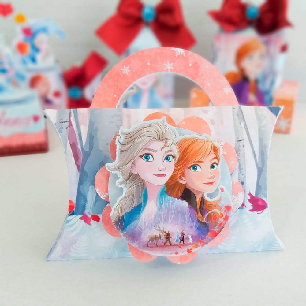 Caixa travesseiro do tema Frozen, formato arredondado com pontas cinchadas e alça arqueada, com medalhão oval recortado com o retrato de Elsa e Anna juntas, fundo com padrão de floresta de inverno e pequenos detalhes de folhas vermelhas