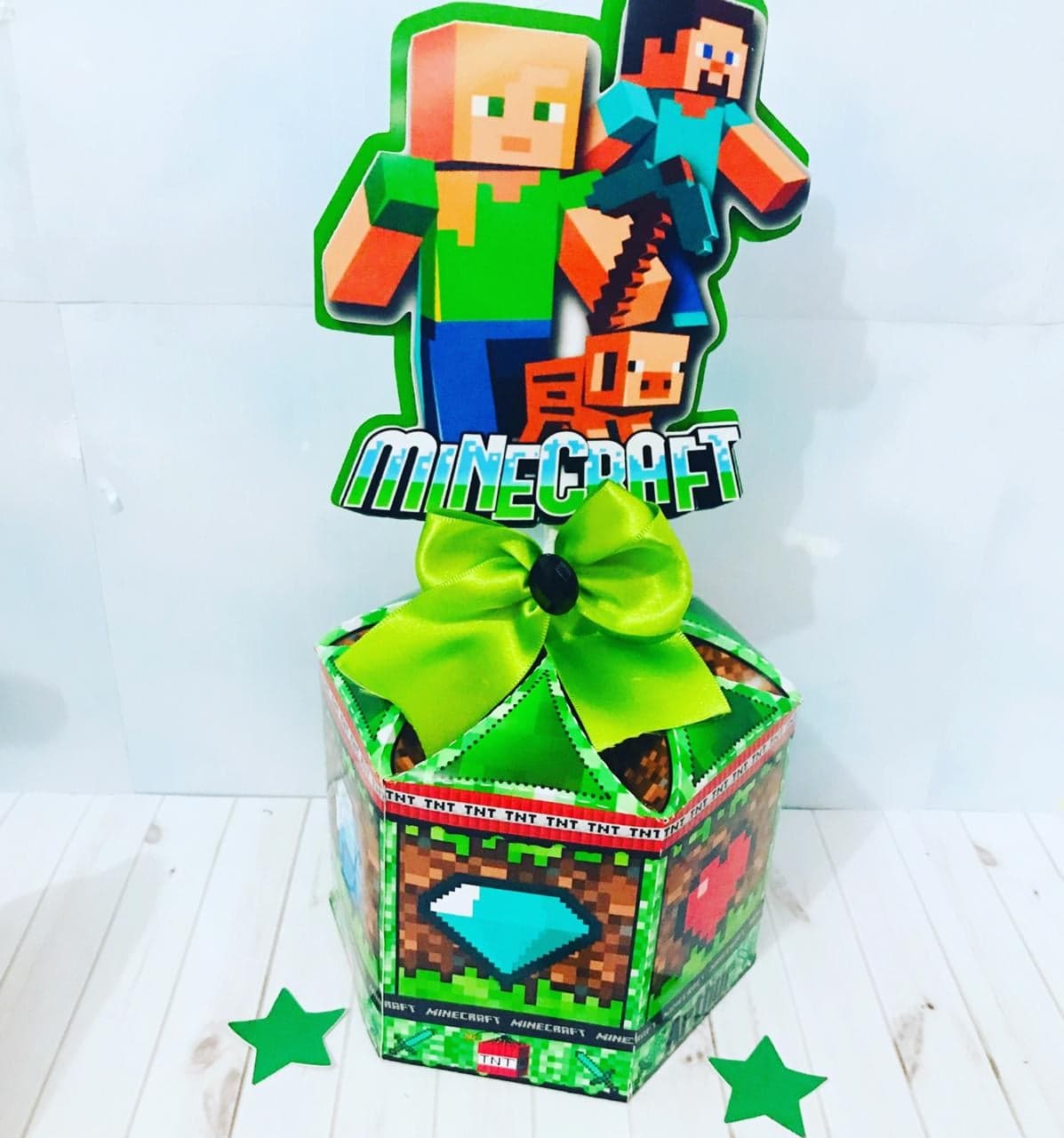 Centro de mesa do tema Minecraft: base hexagonal com textura de bloco e laço verde-limão, coroada por um recorte com os personagens e um porquinho e o logo 'Minecraft'