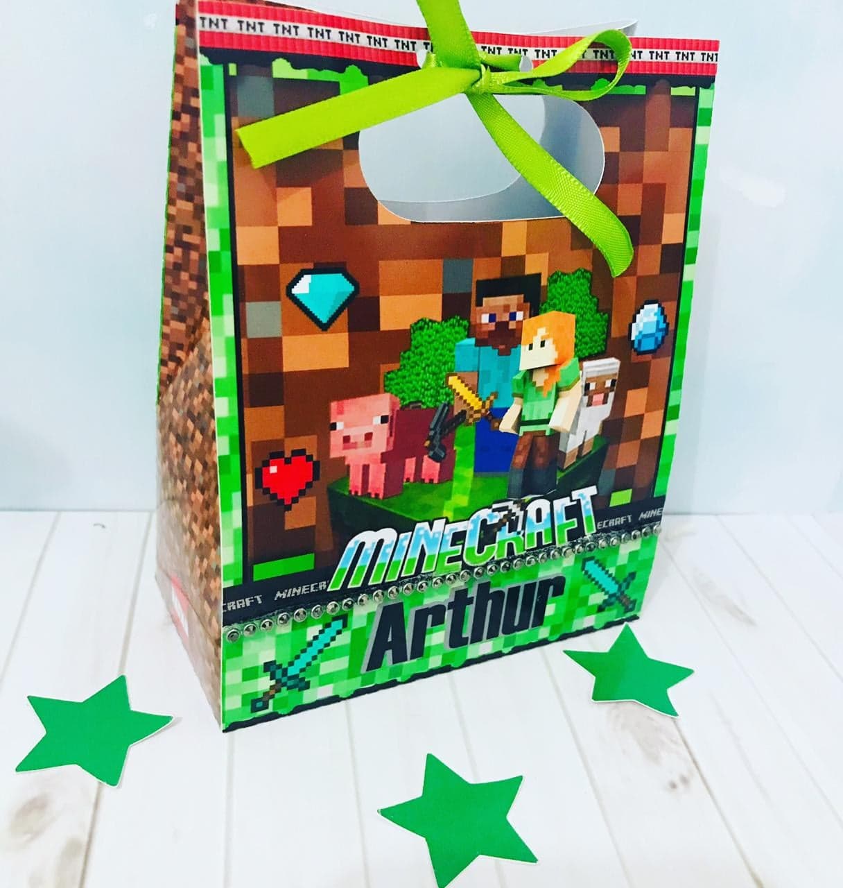 Sacolinha surpresa do tema Minecraft com alça de fita verde-limão, textura de bloco de terra, personagens e animais do jogo, pedrarias na base e o nome 'Arthur'