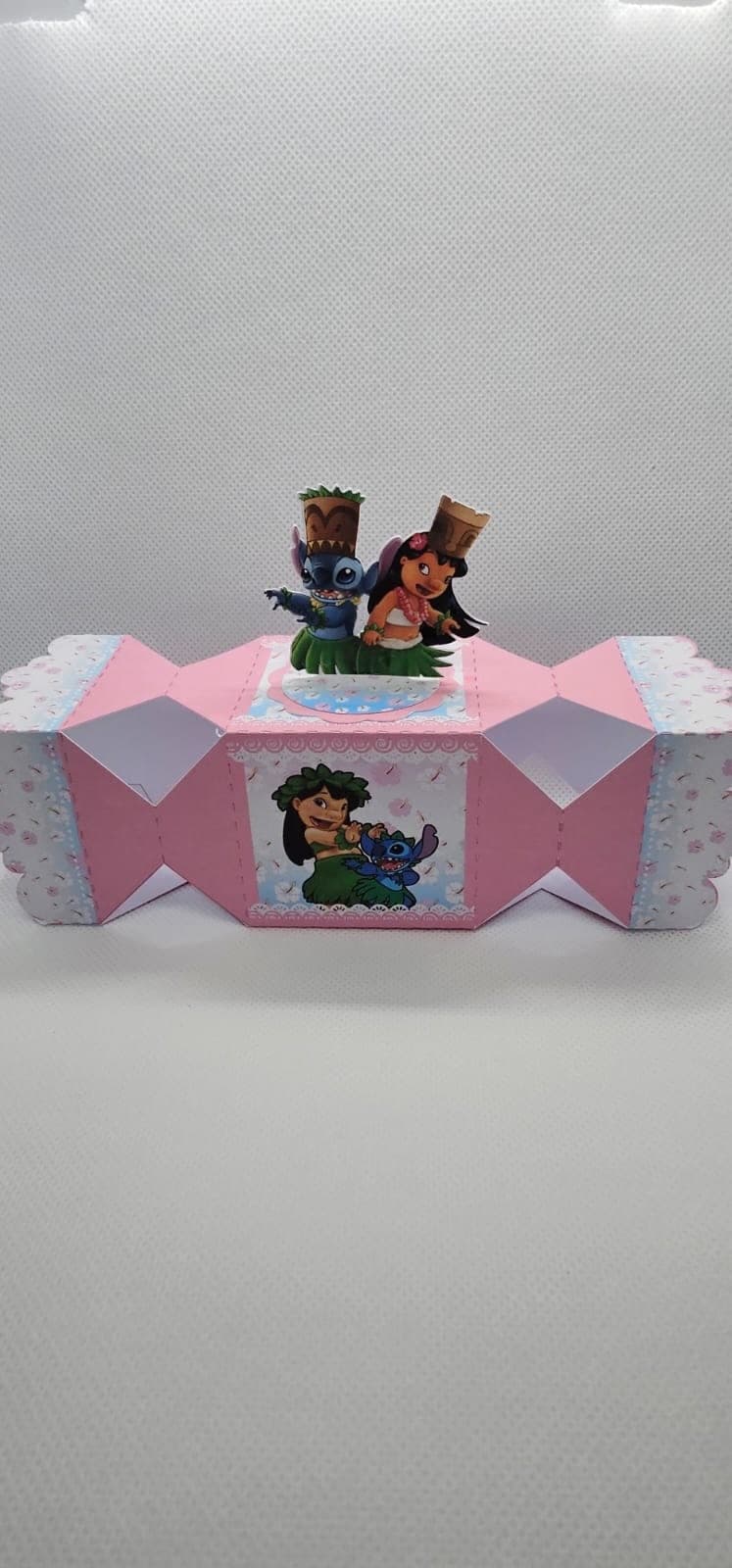 Caixa bala personalizada no tema Stitch, em rosa com estampa floral, coroada por um standee 3D de Lilo e Stitch usando chapéus de casca de coco