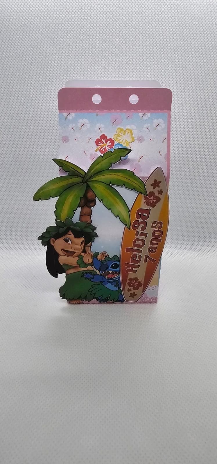 Cartão personalizado do tema Stitch com furos para pendurar, coqueiro de papel, flores havaianas e uma prancha de surfe com o nome 'Heloísa' e a idade